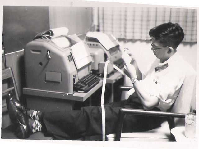 A man in a bow tie feeding punched paper tape through a teleprinter